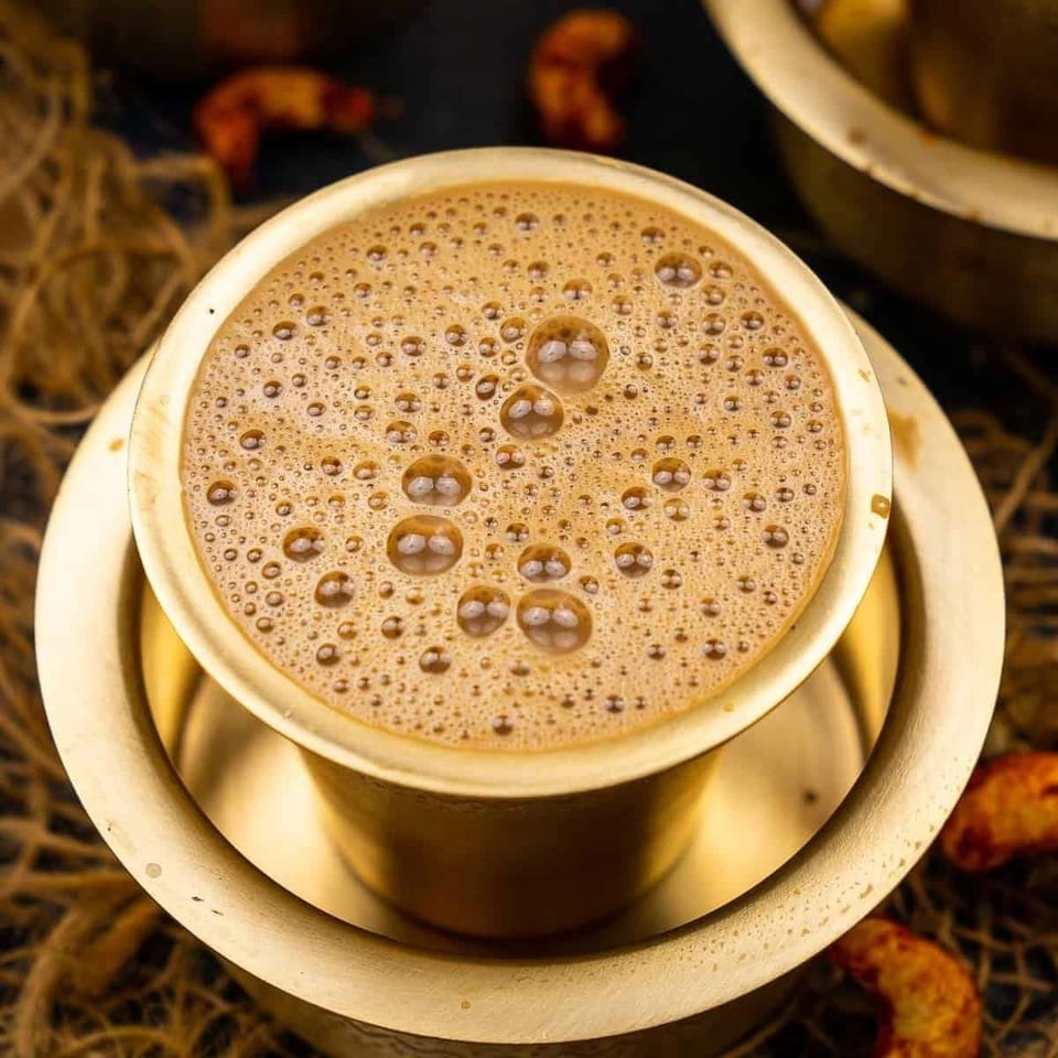 Close-up of a frothy brown beverage served in a cup and saucer, with blurred pastries and woven décor in the background.